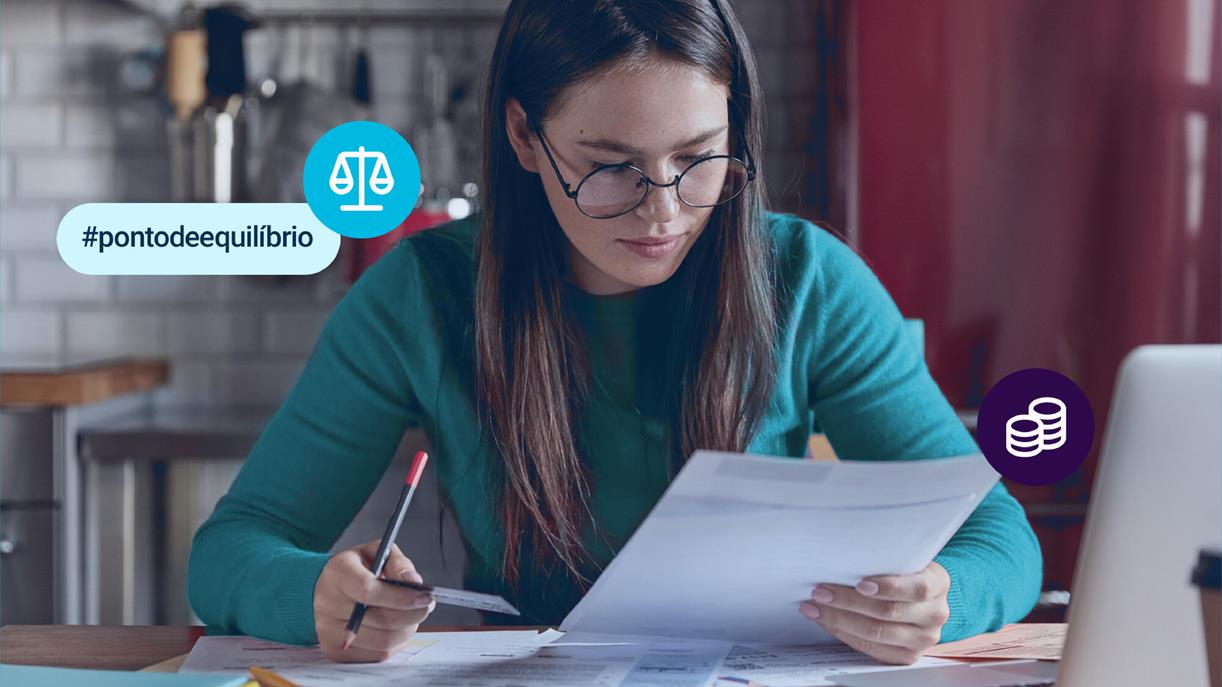 Mulher de óculos sentada à mesa com um laptop e um papel sobre ponto de equilíbrio financeiro.